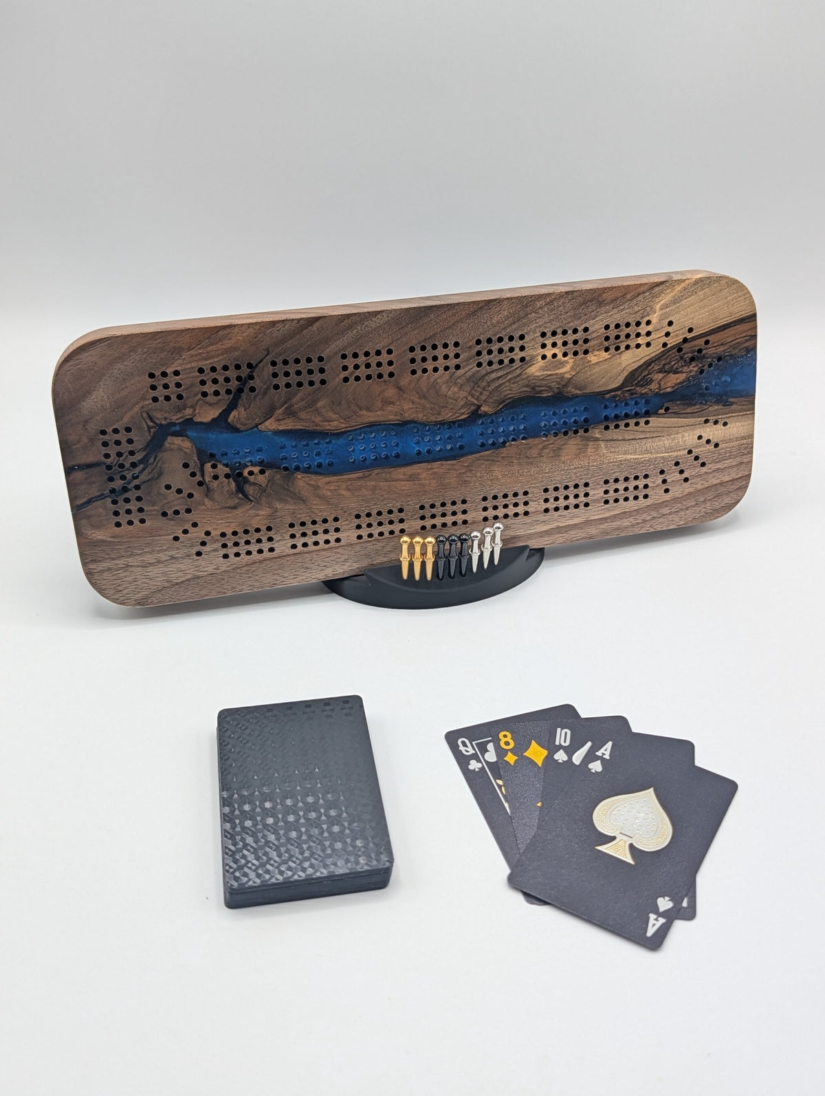 Origin Canadian black walnut cribbage board with a winding cobalt blue epoxy river, displayed with its cards, pegs and stand.